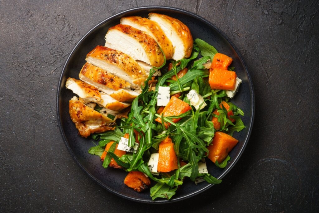 Peito de frango grelhado com salada de rúcula, abóbora e queijo servido em prato preto em cima de bancada de pedra preta