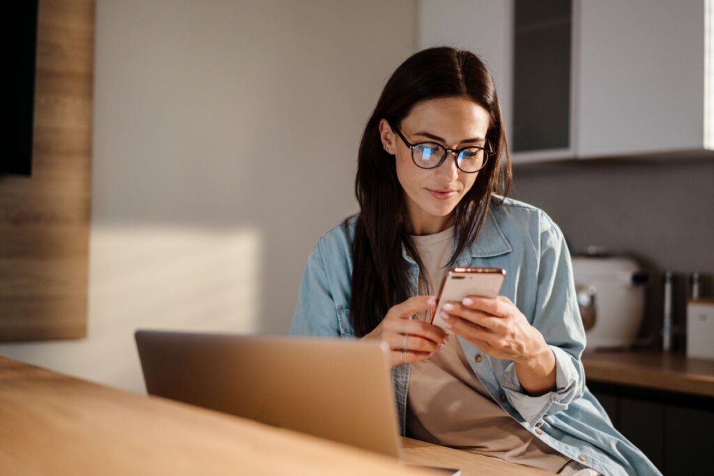 Mulher com cabelo longo, liso e preto, usando camisa de botões jeans e camiseta cinza, óculos preto e com um celular na mão com o notebook ao lado