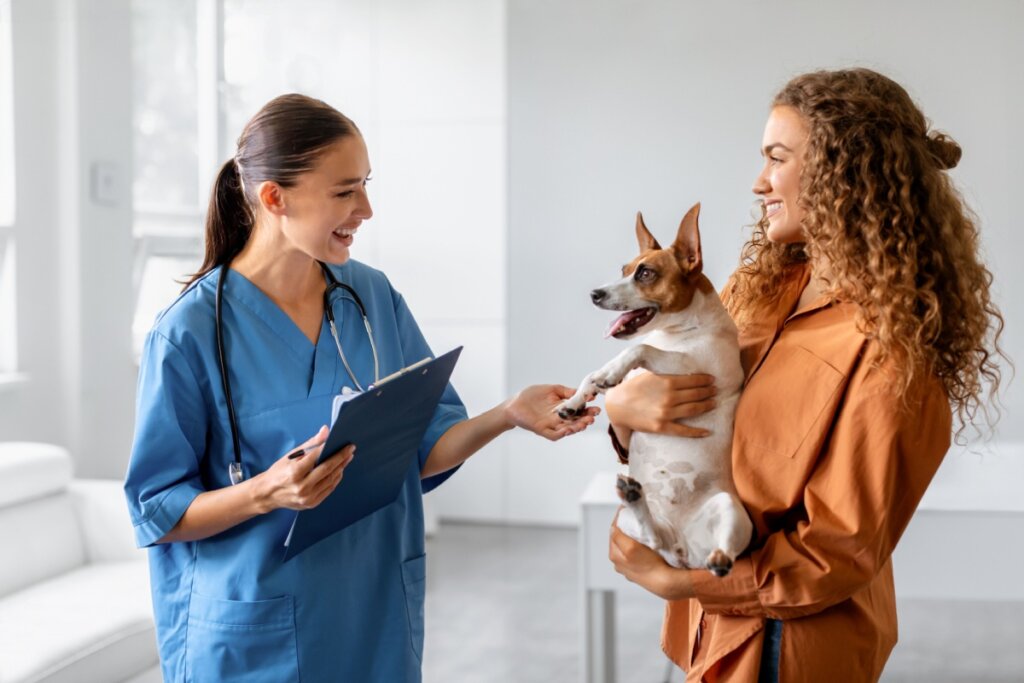 Uma mulher com o cabelo longo amarrado vestida com roupa cirúrgica azul e com uma prancheta na mão e outra mulher com o cabelo solto cacheado e usando uma camisa laranja com um cachorro no colo
