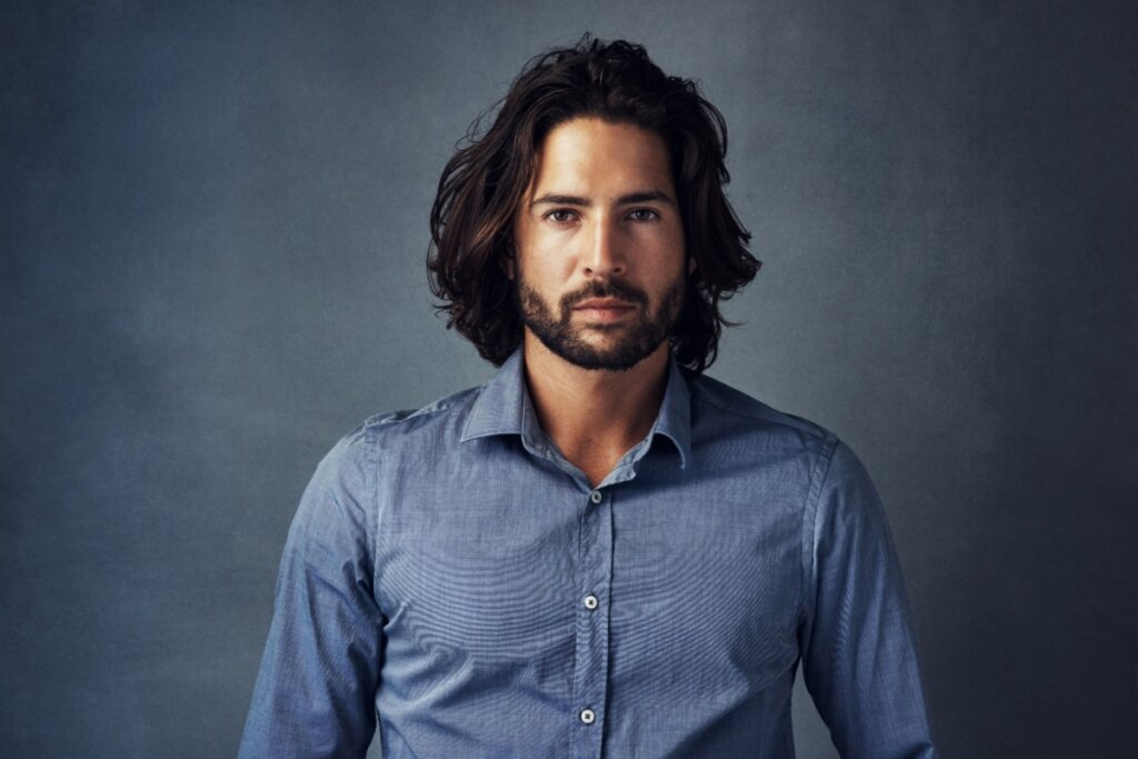 Homem usando camisa azul, parado em frente a um fundo cinza, com o corte longo desconectado e barba média