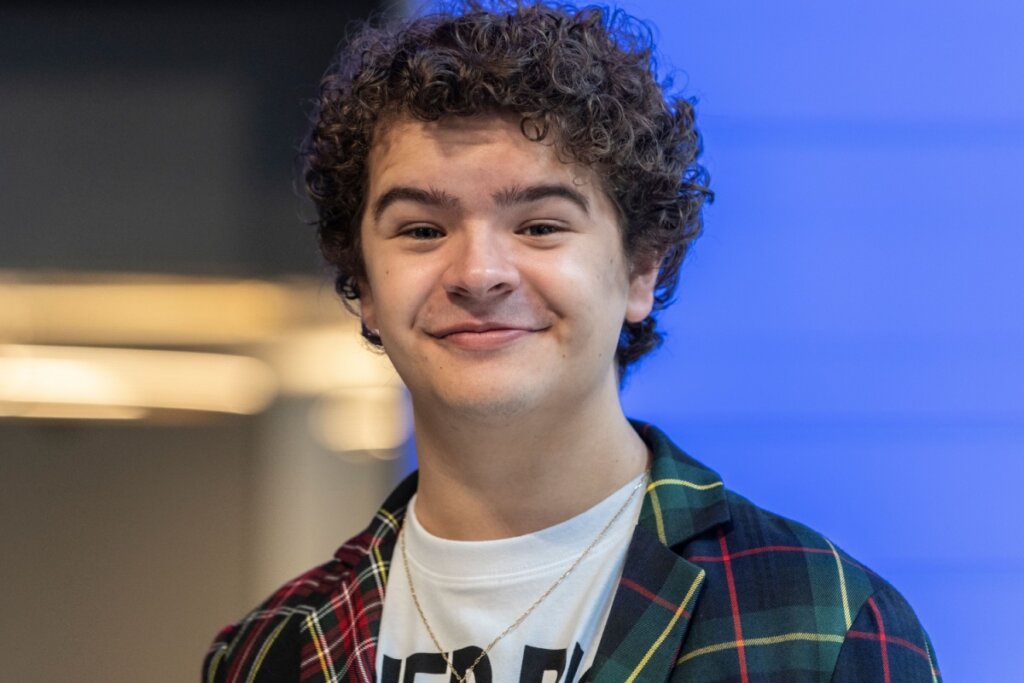 Ator Gaten Matarazzo sorrindo e usando uma blusa xadrez na cor azul com linhas verdes, amarelas e vermelhas