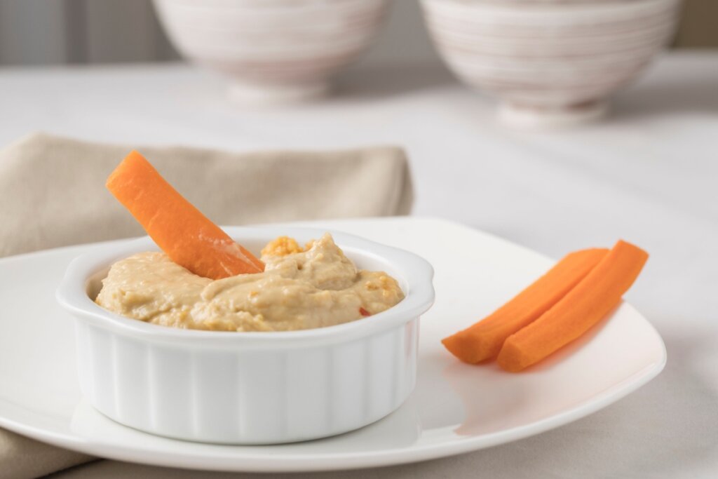 Recipiente branco com homus e um palito de cenouravem cima de um prato branco com dois palitos de cenoura do lado
