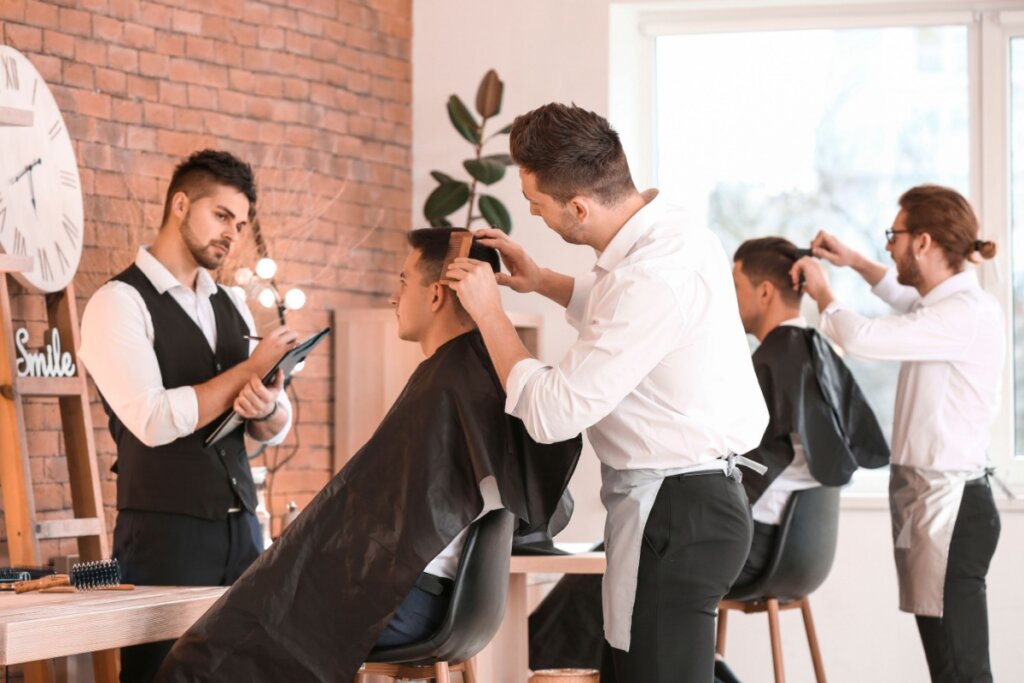 Homens fazendo curso de barbearia e realizando cortes em modelo, com professor acompanhando