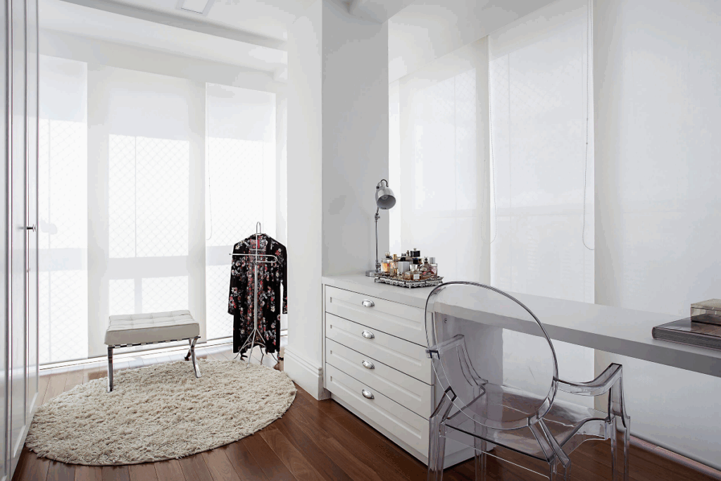 Closet com paredes e móveis brancos, e tapete branco também com parede de vidro e cortina branca e piso de madeira e cadeira em acrílico