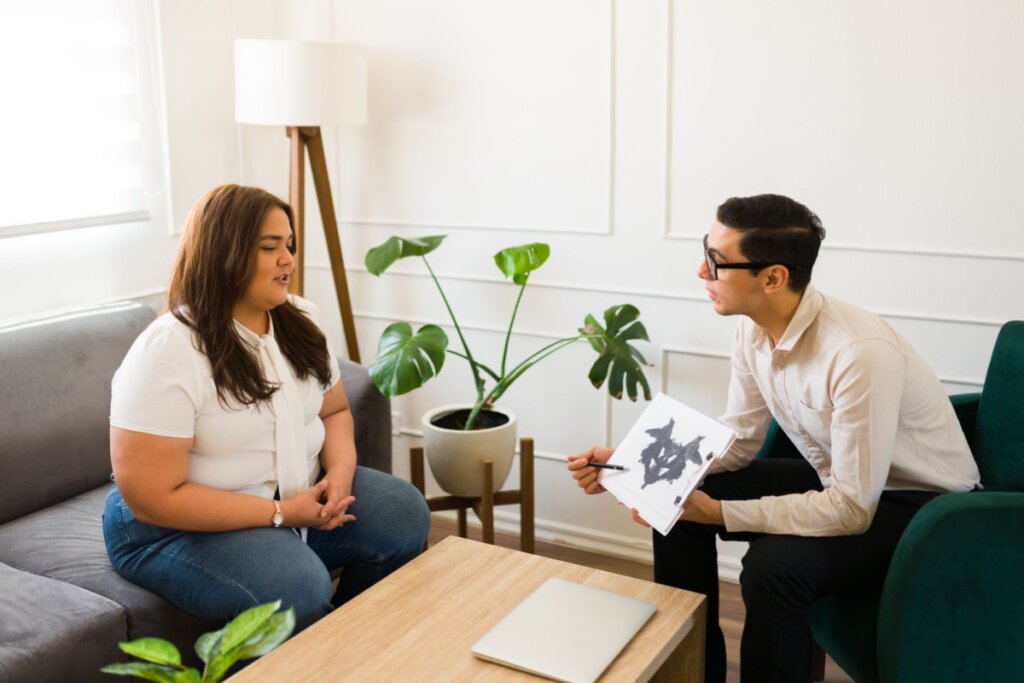 Homem e mulher sentados em sofás separados, frente a frente, enquanto ele mostra a ela um papel com um desenho