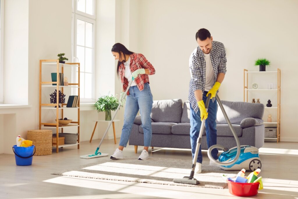 Homem e mulher fazendo faxina em sala de uma casa, com sofá, estantes, tapete e objetos de limpeza ao redor
