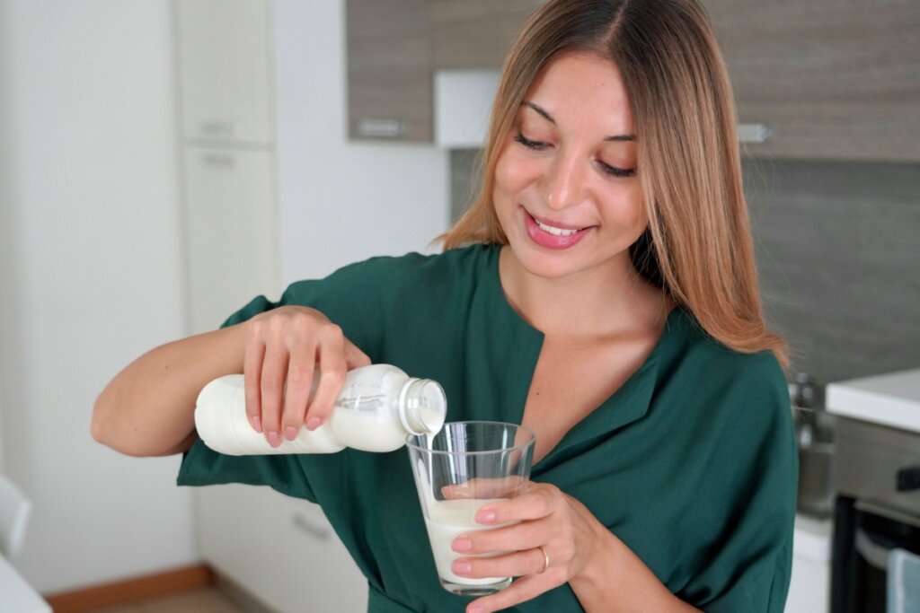 Jovem com cabelo loiro liso, usando camisa verde-escura e colocando leite de kefir em copo para tomar 