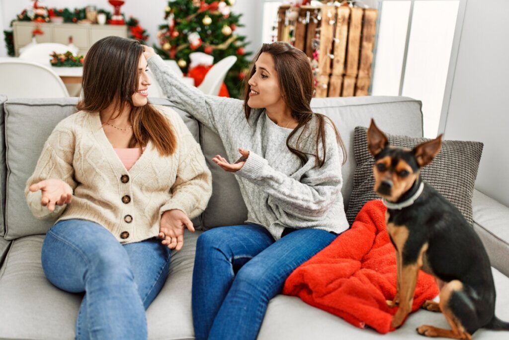 Duas mulheres sentadas em um sofá conversando e sorrindo. Cachorro sentado ao lado 