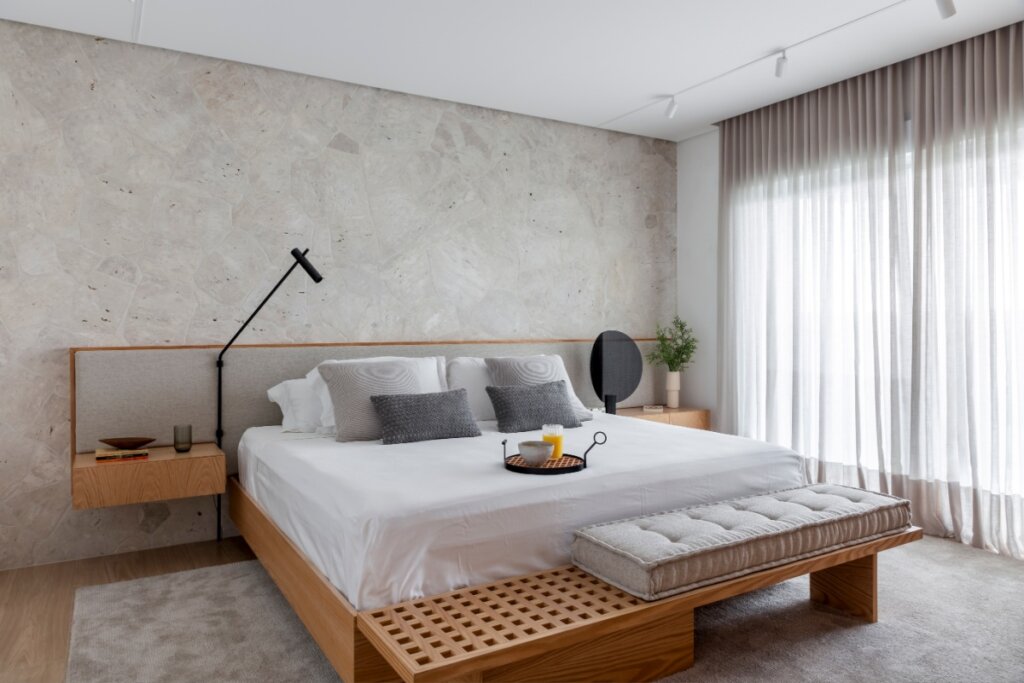 Quarto com cama grande, cabeceira estofada, luminária articulada, banco de madeira e cortina ampla