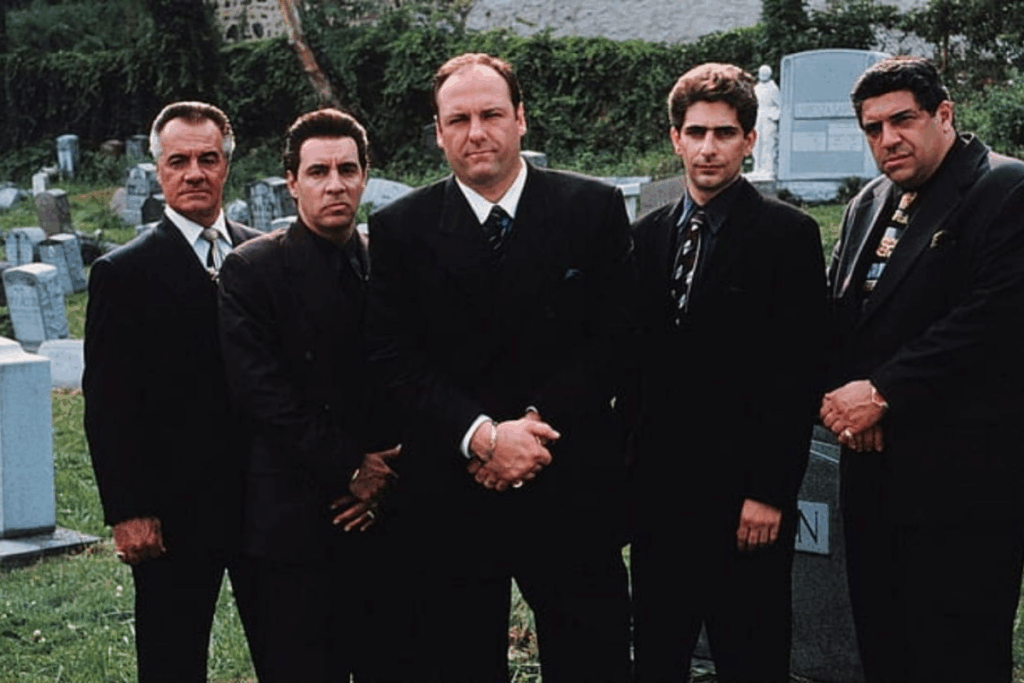 Cinco homens vestidos com ternos escuros e gravatas, alinhados lado a lado, criando uma composição sóbria e intimidadora. Eles estão posicionados de frente para a câmera, com posturas firmes e expressões sérias, transmitindo autoridade