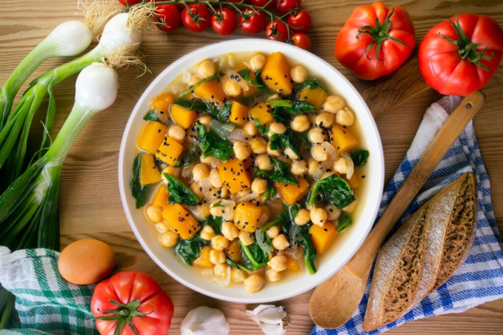 Tigela com grão-de-bico cozido, cubos de abóbora, folhas de espinafre e caldo leve, disposta sobre mesa com legumes e pão ao redor