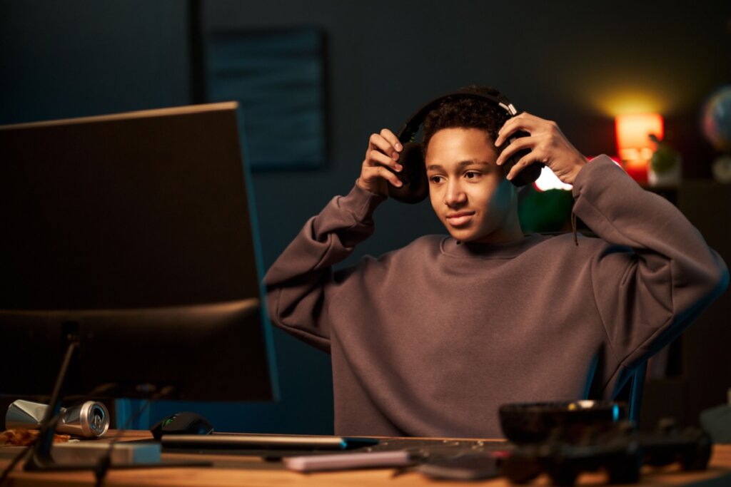 Adolescente negro sentado na escrivaninha colocando fones de ouvido enquanto olha para o monitor do computador, focado em jogos ou atividade on-line no quarto moderno com dispositivos eletrônicos visíveis