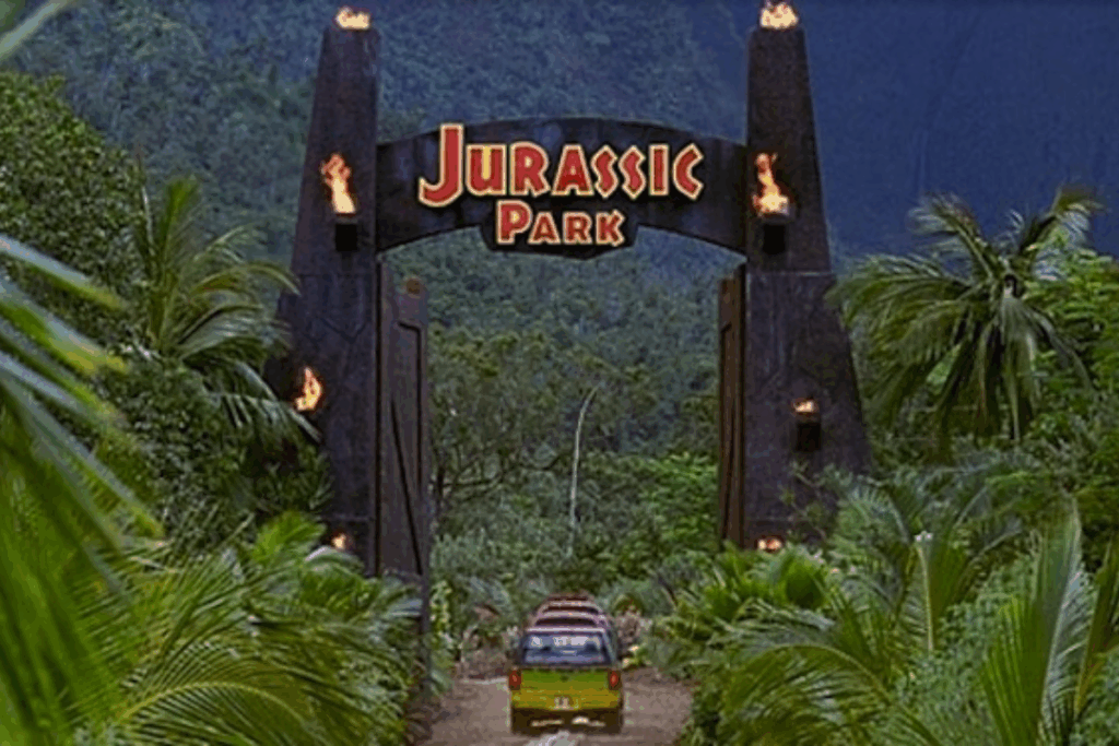 Uma estrada com um carro amarelo e verde na chegada dos portões para Jurassic Park. Ao redor, tem várias árvores e plantas.