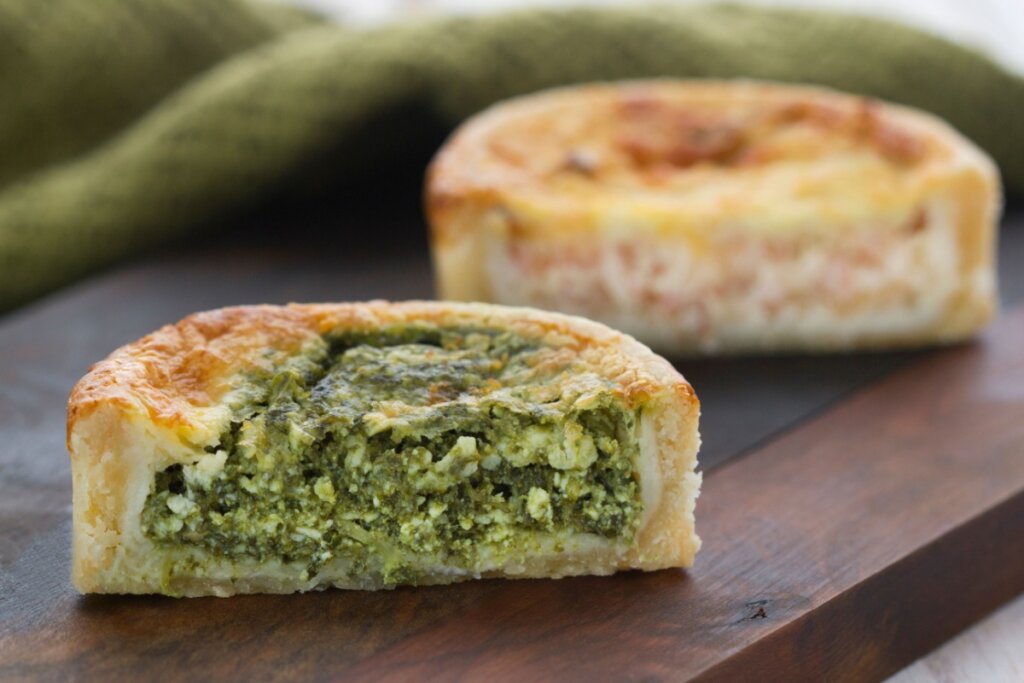 Miniquiche com espinafre e ricota em cima de uma tábua de madeira. Ao fundo, outra miniquiche de frango 