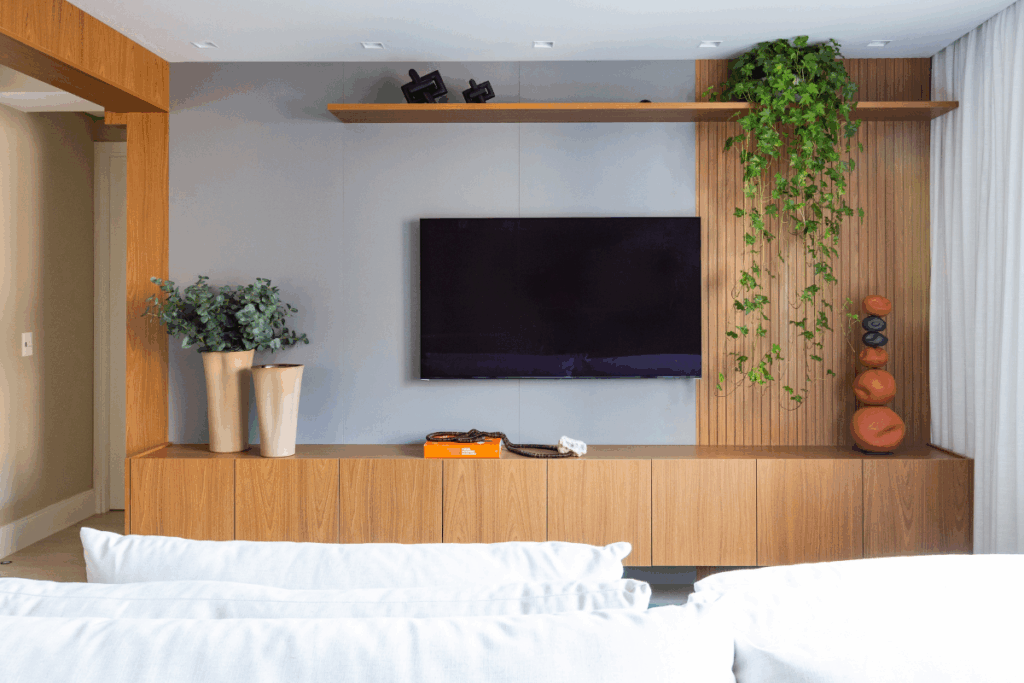 Painel de TV em marcenaria de madeira clara e fundo cinza. Na prateleira superior, à direita, um vaso com uma planta jiboia (ou hera) pendente cai delicadamente sobre o painel ripado de madeira. No móvel inferior, do lado esquerdo, dois vasos altos de cerâmica contêm ramos de eucalipto, criando uma composição equilibrada e orgânica com a tecnologia da TV.