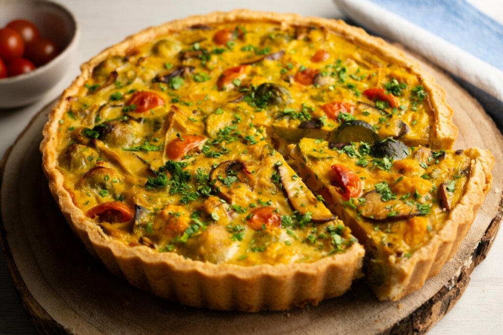 Vista superior de um quiche de batata-doce dourado e rústico, com uma fatia já cortada. O recheio é amarelado e rico, recheado com tomates-cereja e ervas frescas picadas por cima. O prato está sobre uma base de madeira, com uma pequena tigela de tomates ao fundo