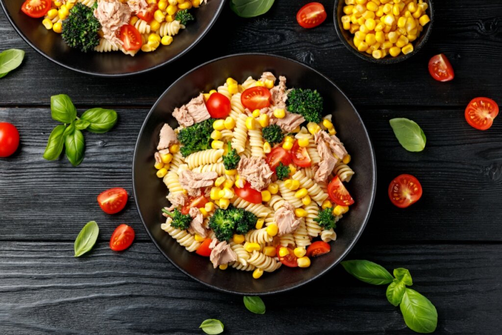 Uma tigela preta vista de cima contendo salada de macarrão fusilli misturada com pedaços de atum, milho-verde, floretes de brócolis cozidos e metades de tomates-cereja