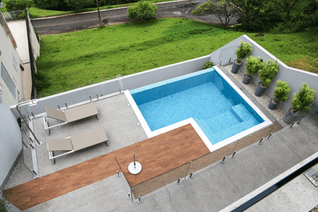 Vista aérea de uma área de lazer externa em um terreno inclinado. Inclui uma piscina retangular com revestimento azul, cercada por um deck de madeira e piso de pedra cinza. Há duas espreguiçadeiras, um chuveiro externo e um guarda-corpo de vidro. Ao fundo, um muro cinza com plantas ornamentais em vasos cinza e uma encosta gramada que leva a uma rua arborizada.