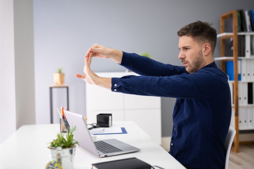 Homem com cabelo curto, usando camisa social azul-escura com os braços pra frente puxando os dedos pra cima, sentado em mesa de escritório