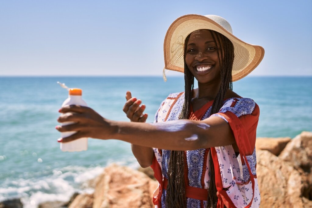 Mulher com cabelo longo, trançado usando chapéu bege e saída de praia vermelha, branca e azul, passando protetor solar no braço