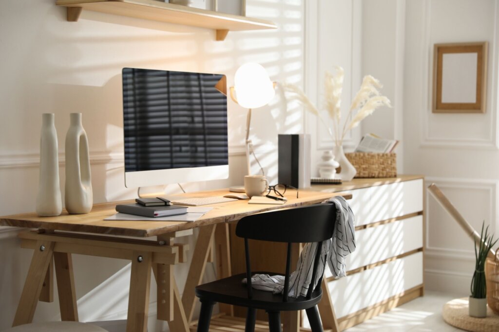 escritório em casa com mesa de madeira e cadeira preta, com móvel com gavetas em madeira e branco ao lado da mesa. Em cima da mesa uma luminária e o monitor do computador com parede clara ao fundo