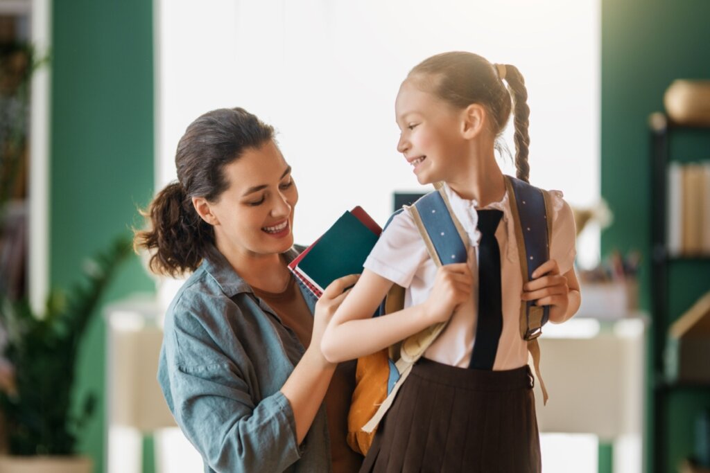 Mulher com o cabelo preso em rabo de cavalo arrumando mochila de menina usando uniforme para ir para a escola