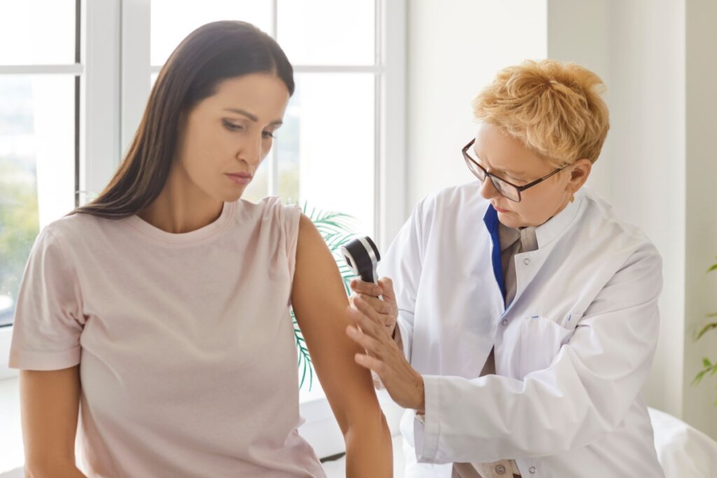 Mulher sendo avaliada por médica em consultório, a paciente tem o cabelo liso e solto e está usando camiseta bege e a médica tem o cabelo loiro curto, está usando jaleco branco e óculos preto, avaliando a pele da mulher