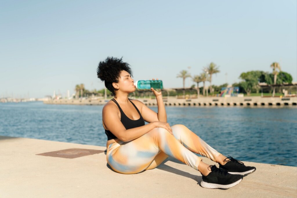 mulher sentada à beira-mar tomando água em garrafa verde. ela usa regata preta, calça colorida e tênis preto