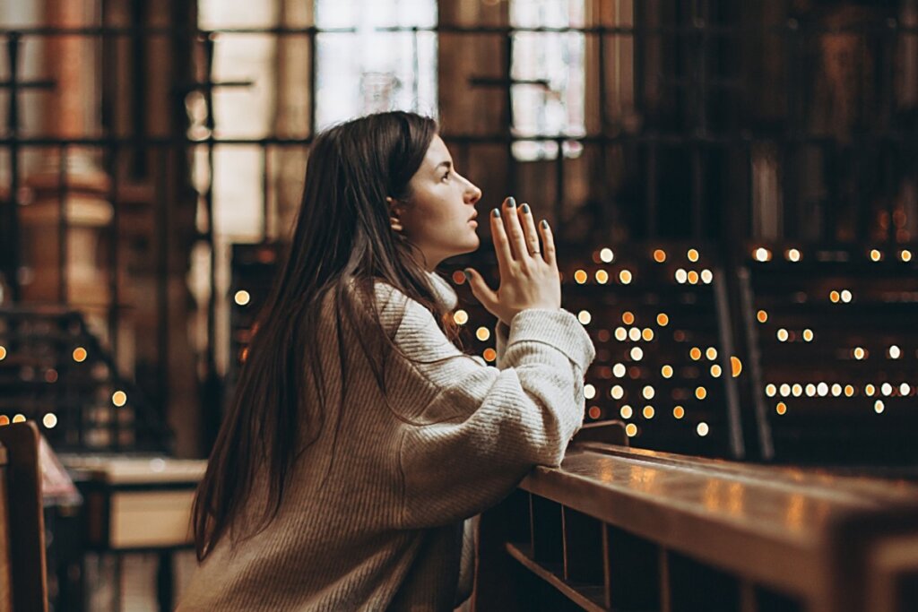 Mulher de cabelos escuros longos vestindo casaco de cor neutra orando em igreja