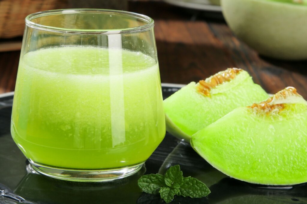 Copo de vidro translúcido com suco verde ao lado de duas fatias de melão