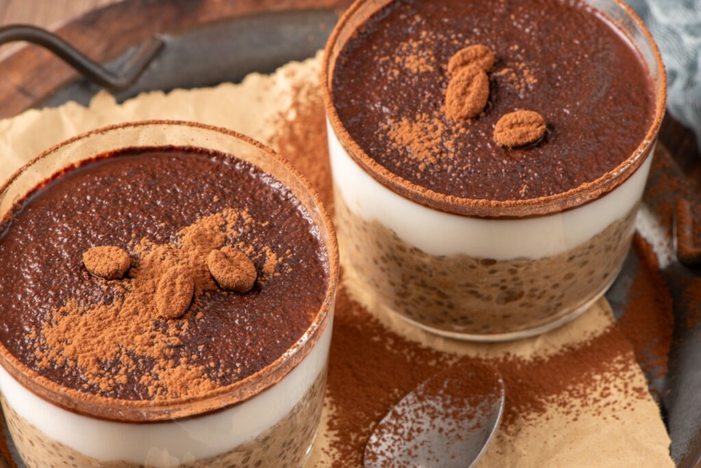 dois copos de vidro com tiramisu com base de chia, creme branco e cacau em pó