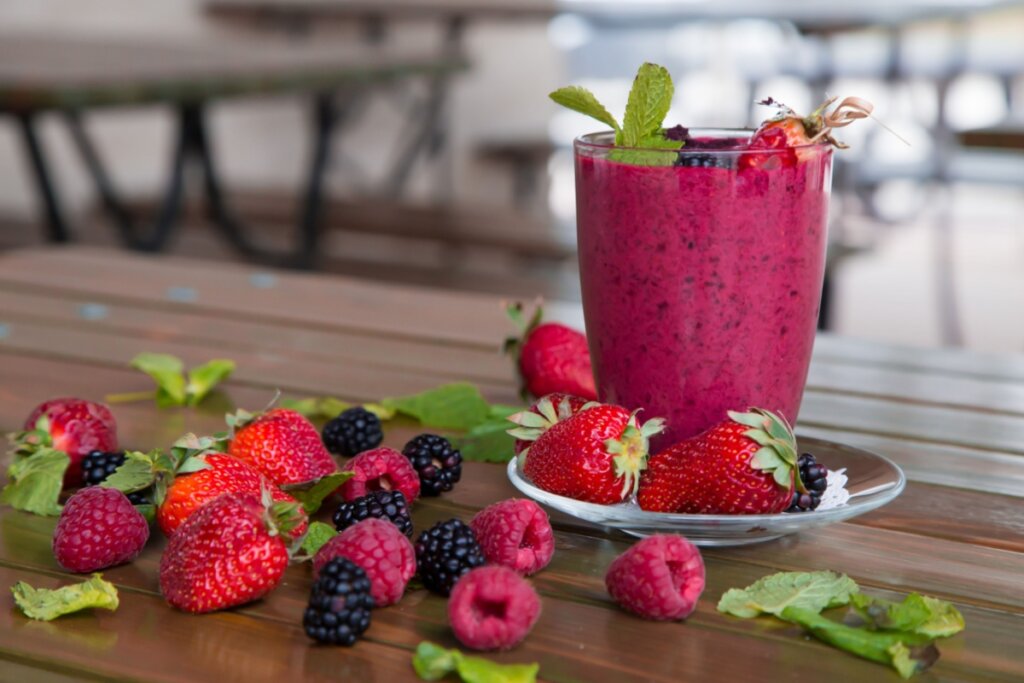 copo de vidro com vitamina vermelha em cima de mesa com frutas vermelhas e folhas verdes em volta
