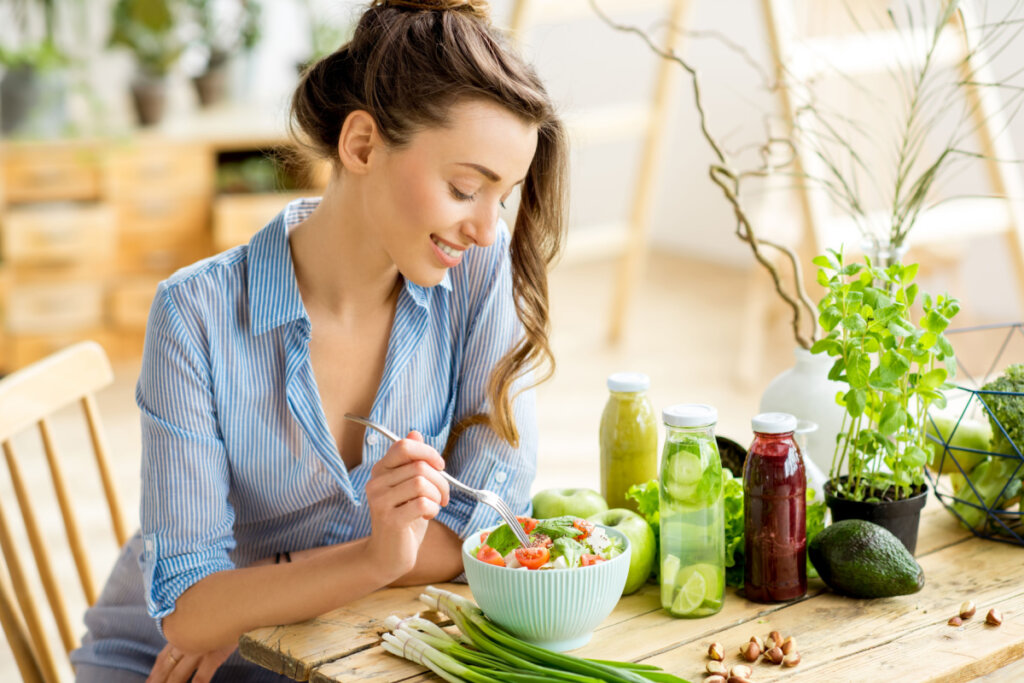 mulher jovem de camisa azul e cabelo preso sentada e com garfo em pote azul com salada com tomate e manjericão. garrafas de suco e frutas em cima da mesa.