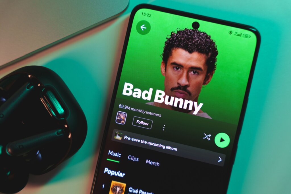 Imagem de um smartphone exibindo o perfil oficial do artista Bad Bunny no aplicativo Spotify. A tela mostra sua foto de perfil com fundo verde vibrante, o nome "Bad Bunny" em letras brancas grandes e a marca de 69,9 milhões de ouvintes mensais. Ao lado do celular, em uma superfície de mesa, aparecem parte de um fone de ouvido preto e um notebook.