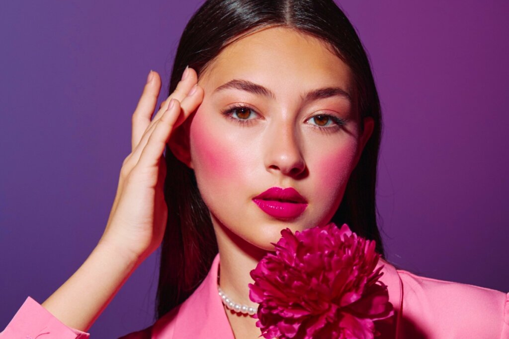 Uma mulher jovem com cabelos castanhos lisos posa em frente a um fundo roxo vibrante. Ela usa uma maquiagem artística monocromática em tons de rosa choque, com um blush intenso e esfumado que se estende das bochechas até as têmporas e pálpebras. Seus lábios estão pintados com um batom rosa brilhante. Ela veste um blazer rosa, usa um colar de pérolas e segura uma flor peônia de cor fúcsia próxima ao rosto, com uma das mãos tocando levemente a lateral da testa.