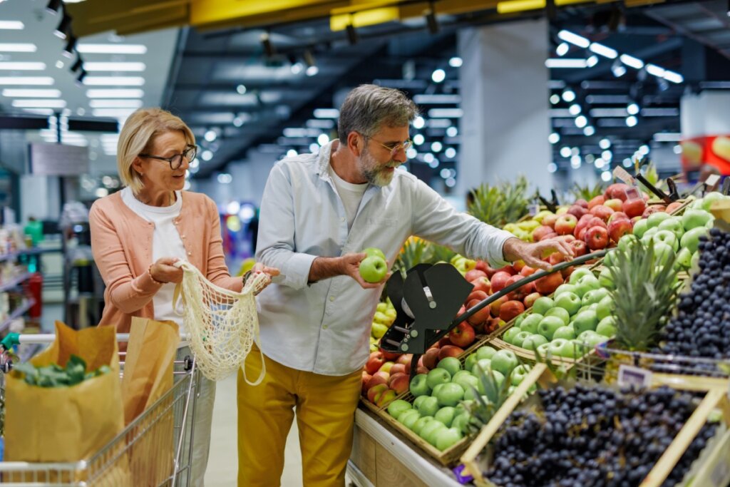 Um casal de meia-idade faz compras na seção de hortifrúti de um supermercado iluminado. O homem, com barba grisalha e camisa clara, segura uma maçã verde enquanto escolhe outras frutas em uma bancada repleta de maçãs vermelhas, verdes e uvas. Ao lado dele, a mulher, de óculos e cardigã rosa, segura uma sacola de rede reutilizável aberta, pronta para guardar as frutas. À frente deles, há um carrinho de compras com sacos de papel cheios de mantimentos.