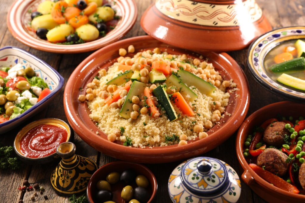 Prato de cuscuz marroquino servido em travessa de cerâmica, acompanhado de legumes variados e grão-de-bico, com molhos e acompanhamentos ao redor
