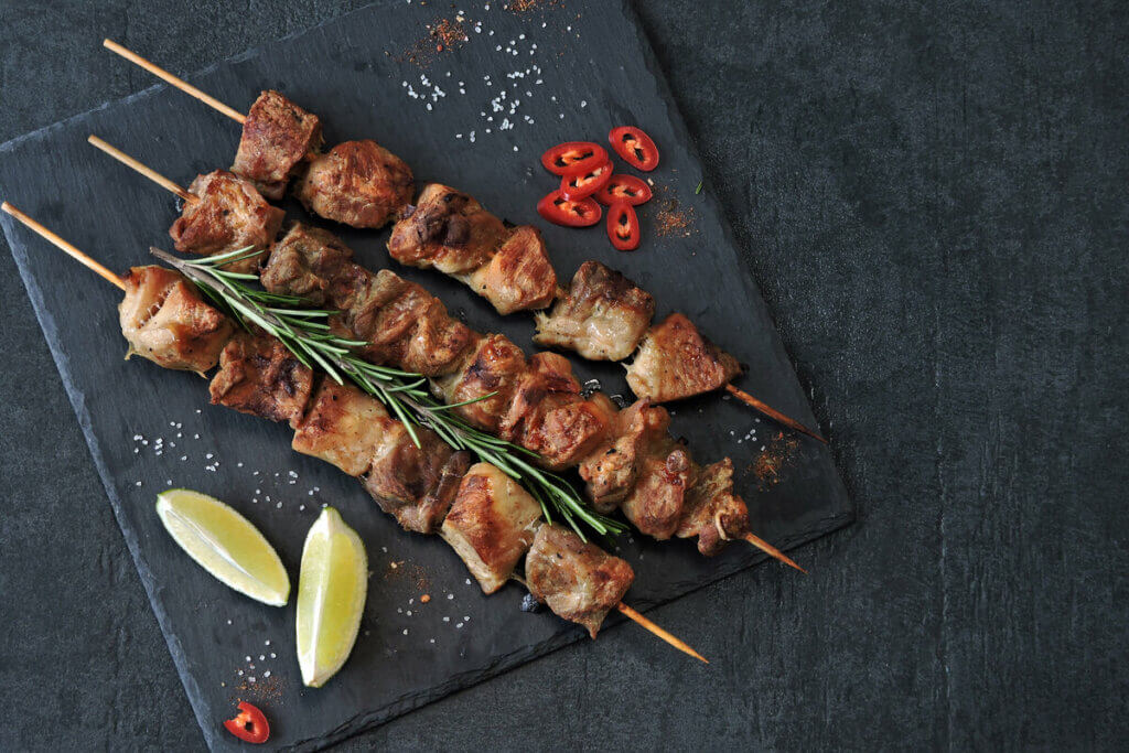 Espetinhos de carne grelhada servidos sobre uma tábua escura, acompanhados de fatias de limão, pimenta vermelha em rodelas, sal grosso e um ramo de alecrim