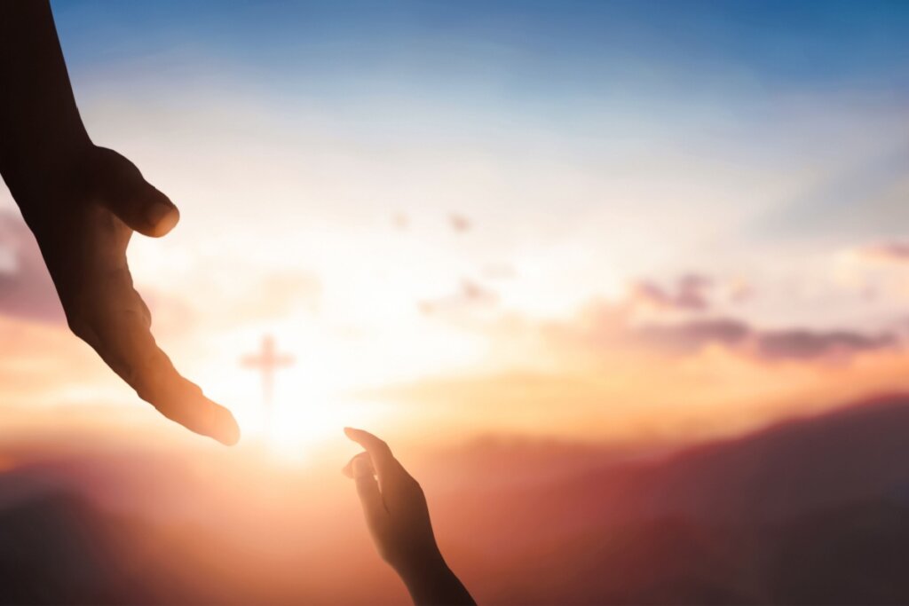 Silhueta de duas mãos, uma maior descendo e outra menor subindo, quase se tocando contra um pôr do sol vibrante. Ao fundo, uma cruz cristã está centralizada sob a luz do sol entre montanhas.