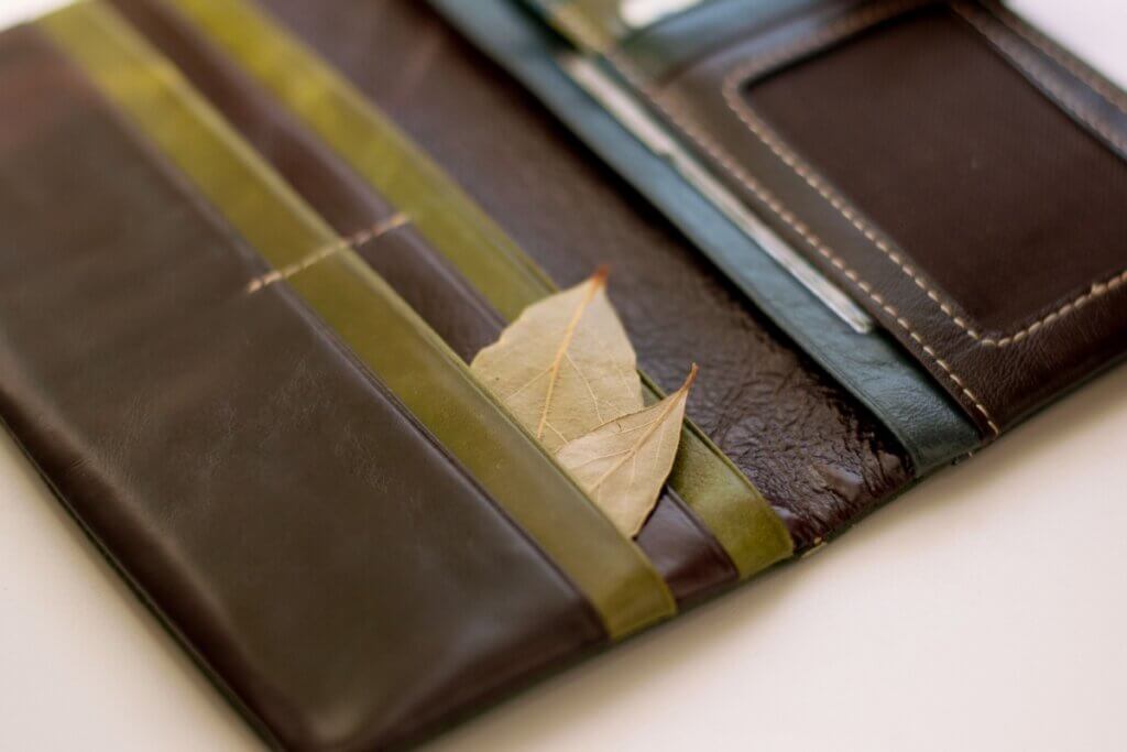 Uma carteira de couro aberta, em tons de marrom e verde-oliva, sobre uma superfície clara. Dentro de um dos compartimentos destinados a cartões, estão inseridas duas folhas de louro secas