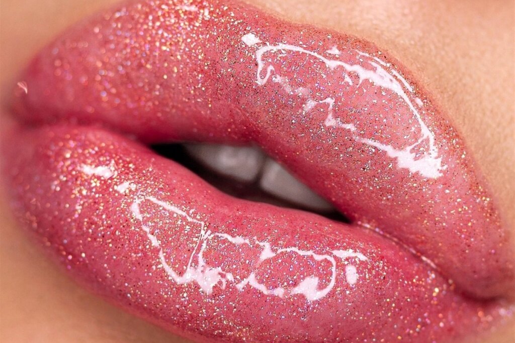 Fotografia em alta definição de lábios femininos entreabertos. Eles estão cobertos por um batom rosa cintilante repleto de micropartículas de glitter multicolorido. Por cima, uma camada de gloss espelhado reflete luzes brilhantes, realçando a textura granulada do glitter e o volume dos lábios. É possível ver detalhes dos dentes brancos ao fundo.