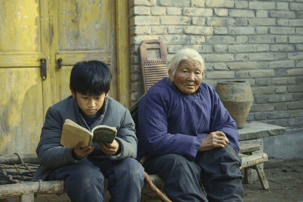 Um menino e uma idosa de etnia asiática estão sentados lado a lado em um banco de madeira rústico à frente de uma parede de tijolos claros. O menino, à esquerda, veste um casaco azul-escuro e lê atentamente um livro pequeno. A idosa, à direita, possui cabelos brancos presos e veste uma túnica azul acolchoada, olhando diretamente para a câmera com uma expressão serena e marcada pelo tempo. Uma porta amarela desgastada compõe o cenário ao fundo.