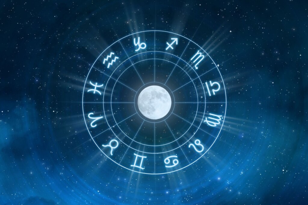 Ilustração digital de um mapa astrológico sobreposto a um fundo de céu noturno estrelado em tons de azul profundo e nebulosas sutis. No centro exato da imagem, há uma lua cheia brilhante e detalhada. Ao redor da lua, um anel circular dividido em doze segmentos contém os símbolos brancos e brilhantes do zodíaco (como Áries, Touro, Gêmeos, entre outros). Feixes de luz azul clara emanam do centro em direção às bordas, criando um efeito de brilho e expansão mística em toda a composição.