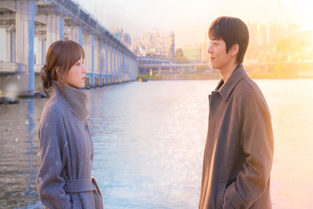 Homem e mulher se encarando à beira de um rio, com ponte e prédios ao fundo sob luz suave