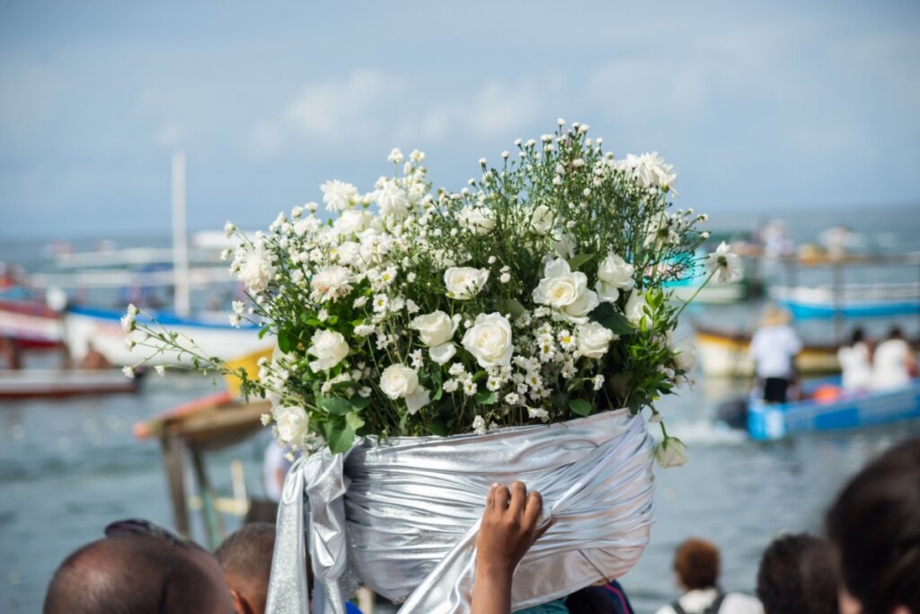 Fotografia colorida em plano médio capturada no Rio Vermelho, Salvador. Em destaque, uma pessoa carrega sobre a cabeça um grande e vistoso cesto de oferendas para Iemanjá, envolto em um tecido prateado brilhante e acetinado. O cesto transborda com um arranjo denso de flores brancas, incluindo rosas e pequenas flores de campo (mosquitinho), simbolizando pureza e devoção. Ao fundo, o mar da Bahia está repleto de barcos coloridos e fiéis, sob um céu levemente nublado, durante as celebrações do dia 2 de fevereiro