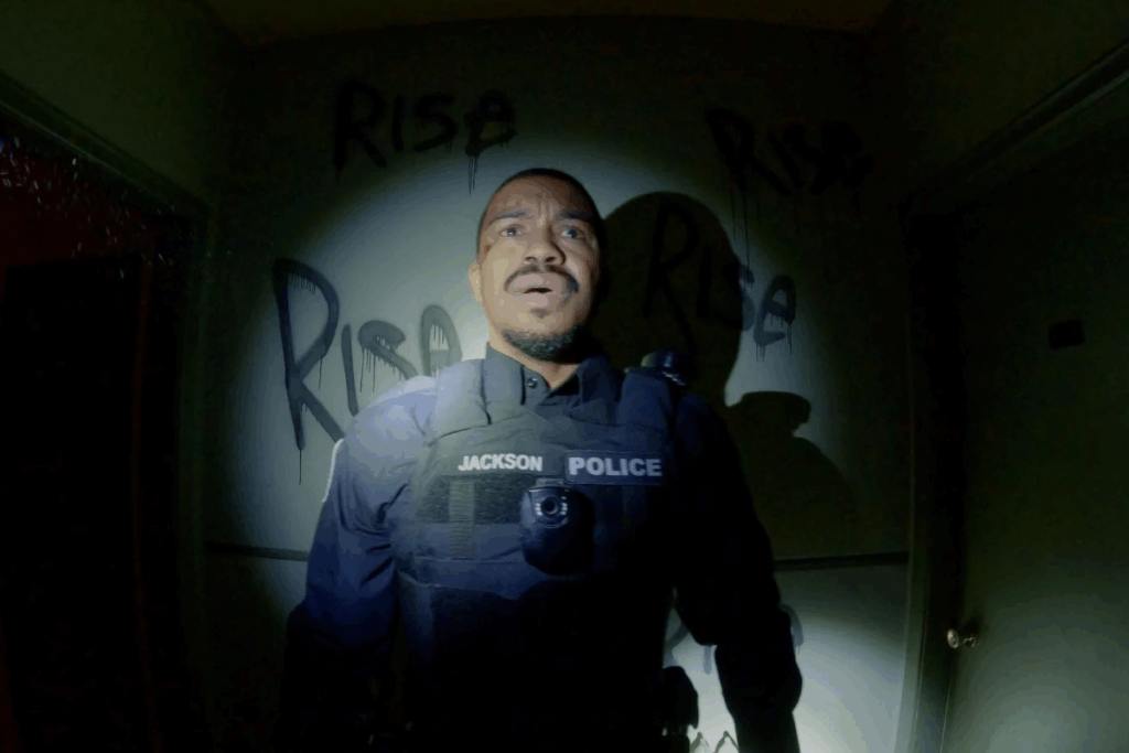 Uma imagem escura capturada de uma perspectiva em primeira pessoa (estilo "bodycam"). O foco está em um policial negro, cujo colete exibe os dizeres "JACKSON POLICE". Ele parece ofegante ou surpreso, iluminado por uma lanterna direta. Na parede atrás dele, há pichações com a palavra "RISE" escritas de forma agressiva. O ambiente é um corredor estreito e sombrio, transmitindo uma atmosfera de suspense ou terror.