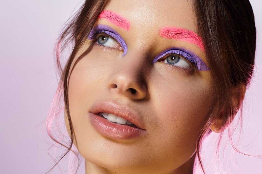 Rosto de uma mulher jovem com uma maquiagem artística e colorida. Ela olha para cima e para a esquerda, revelando sombras em tom lilás vibrante aplicadas de forma gráfica nas pálpebras superiores. O destaque principal são as sobrancelhas, que foram totalmente cobertas por uma cor rosa neon intensa com textura visível. A pele tem um acabamento natural e iluminado, os lábios estão com um gloss suave e fios de cabelo castanho emolduram o rosto sobre um fundo lilás claro.