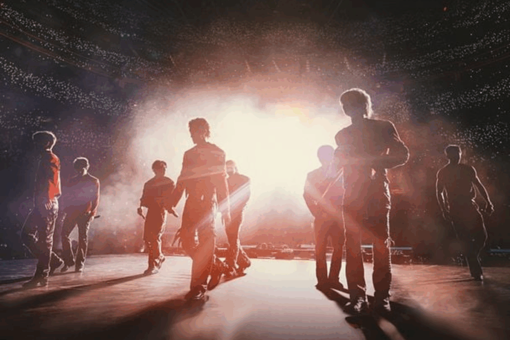 Fotografia de um show musical em um grande estádio lotado. Os integrantes do grupo Stray Kids aparecem como silhuetas contra uma luz branca central extremamente forte e radiante que emana do palco, criando um efeito de feixes de luz.