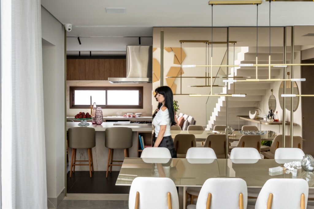 mpla sala de jantar integrada com a cozinha ao fundo. Em primeiro plano, uma mesa de jantar com cadeiras brancas e um pendente dourado moderno e linear. Uma divisória de vidro reflecta separa sutilmente os ambientes, refletindo a escada de design minimalista. Uma mulher caminha pelo espaço.