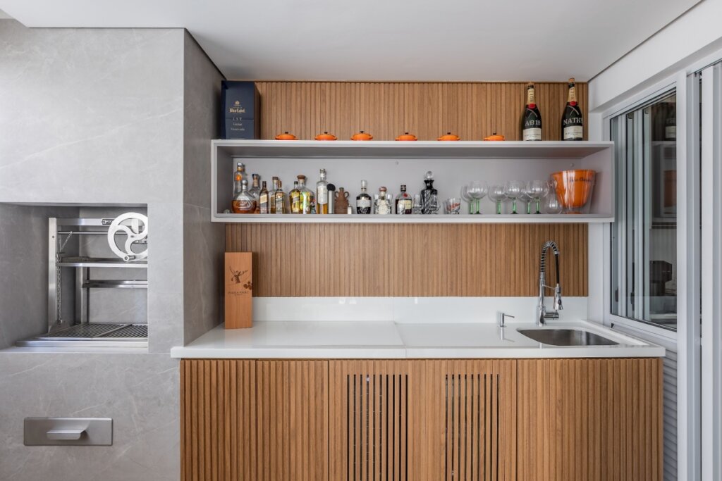 Varanda gourmet com bancada, nicho de bar acima da pia e prateleiras com bebidas e utensílios